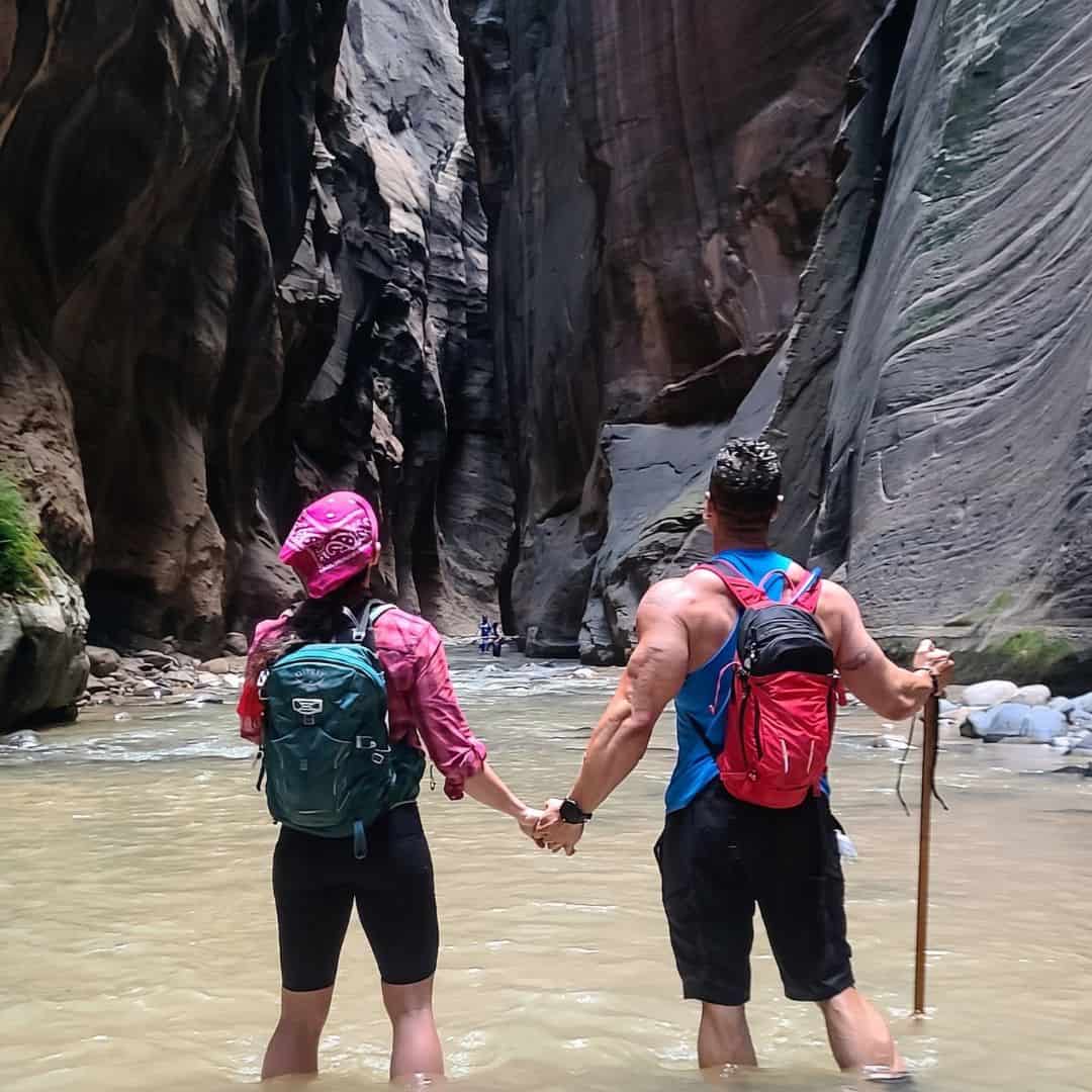 Couple hiking through narrow slot canyon with backpacks and walking stick.
